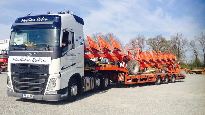 Un camion transportant plusieurs machines agricoles orange, garé dans une aire ouverte, avec des arbres en arrière-plan et un ciel partiellement nuageux.