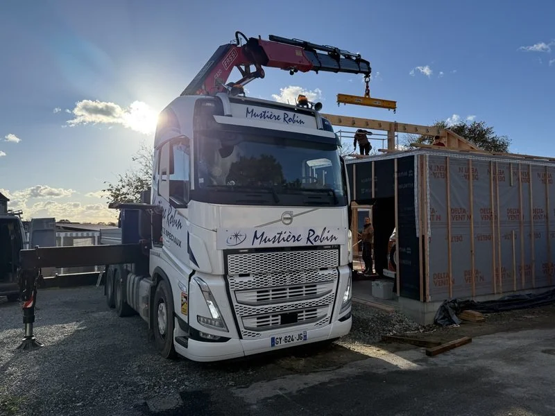 Un camion de construction avec une grue déplaçant une poutre en bois pour une maison en cours de construction