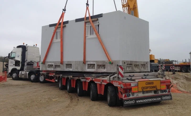 Une grande structure en béton avec des fenêtres, suspendue par des sangles sur un camion de transport sur un site de construction.