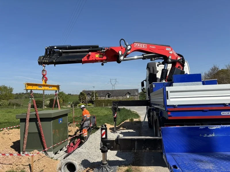 Une machine de levage rouge avec une grue est en train de manipuler une boîte électrique verte sur un chantier en plein air, avec deux ouvriers en travaux et un environnement rural en arrière-plan.