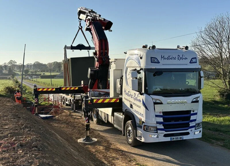 Un camion équipé d'une grue effectue des travaux de construction ou de levage sur le bord de la route, avec deux ouvriers à proximité et un paysage rural en arrière-plan.