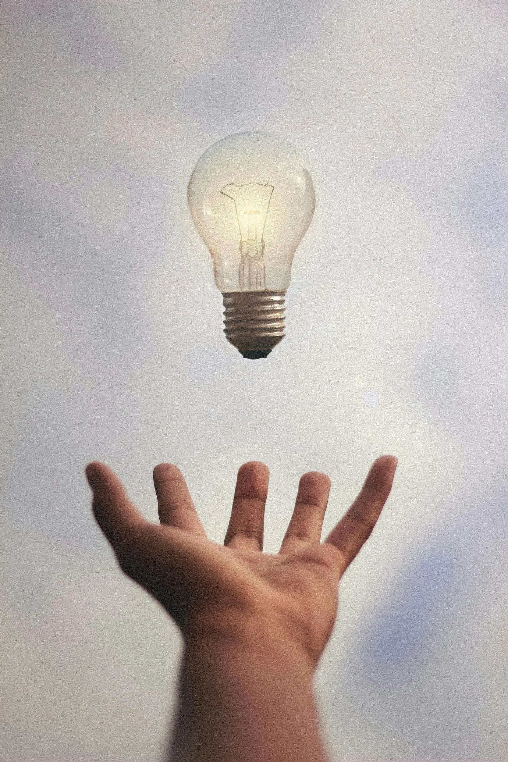 A person's hand with fingers spread, reaching towards a floating lightbulb against a plain white background.