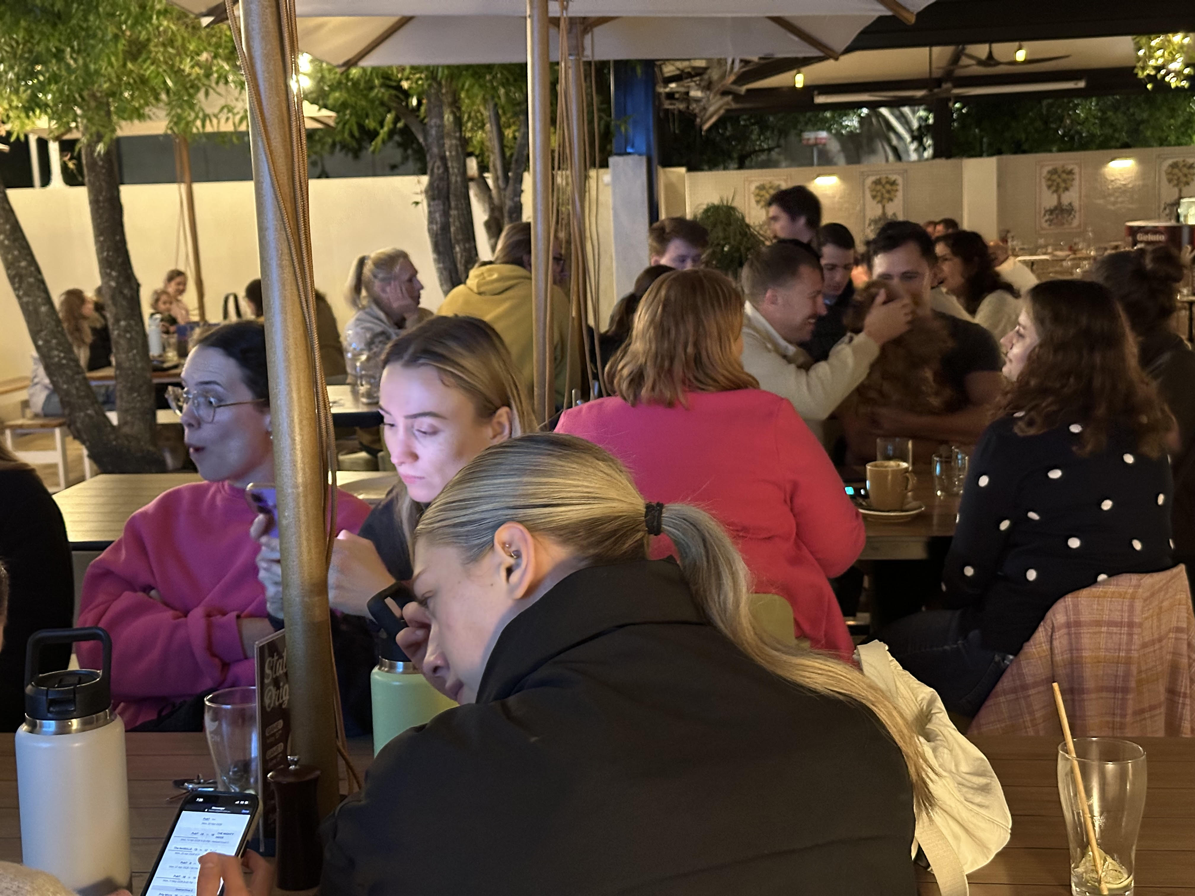 A crowded outdoor restaurant or café at night with people sitting at tables, some using their phones, surrounded by trees and lit with warm lighting.