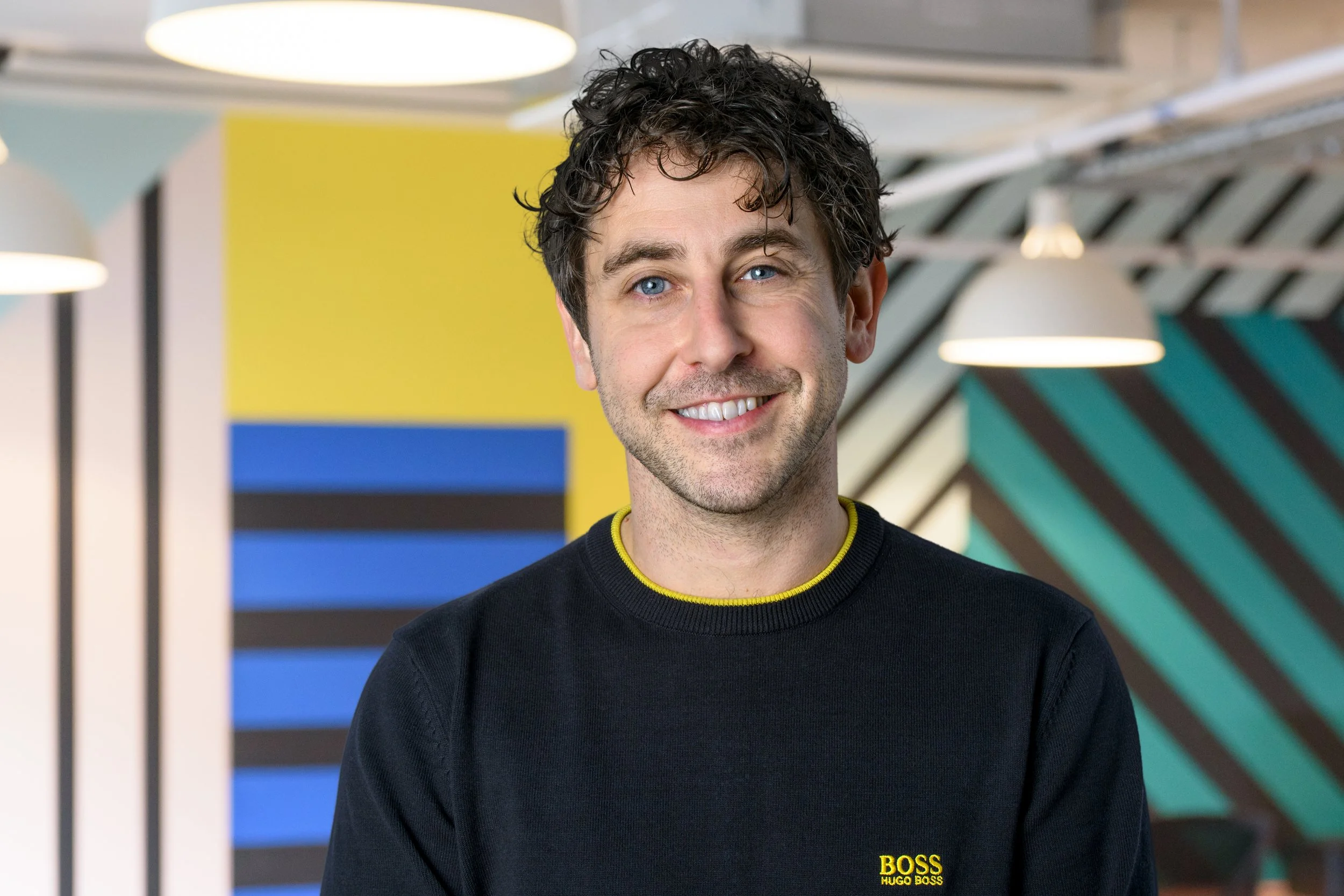 A young man with curly dark hair and blue eyes smiling, wearing a black sweatshirt with yellow collar and yellow "BOSS Hugo Boss" logo, standing in a colorful modern interior with yellow, blue, black, and teal geometric wall patterns and hanging light fixtures.
