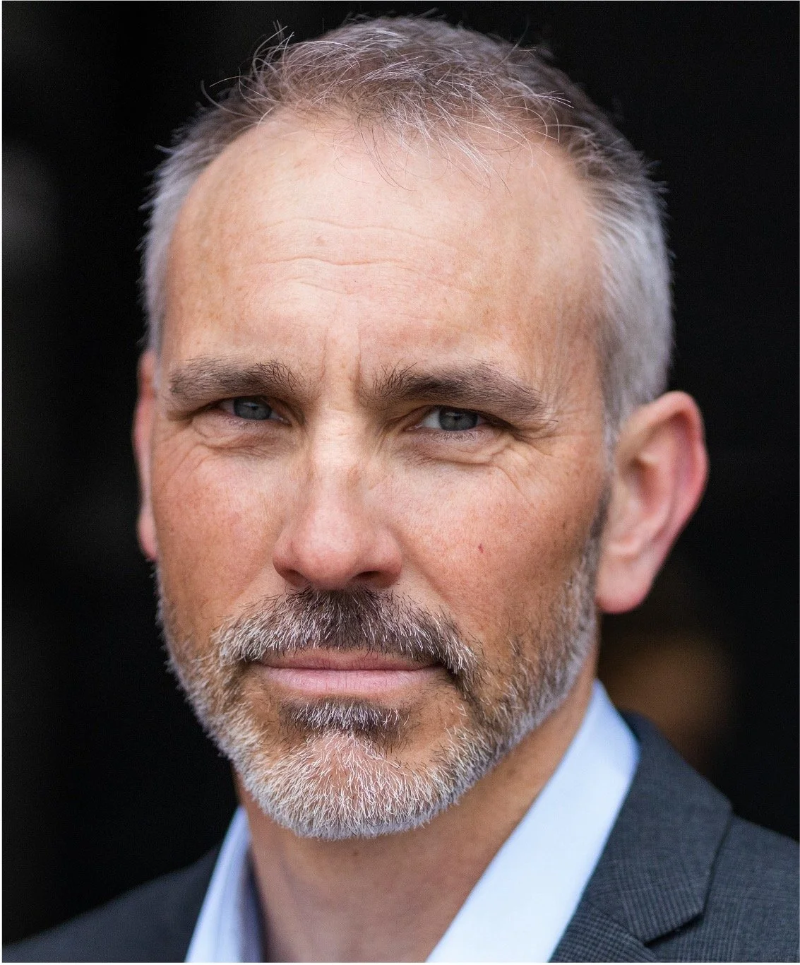 Close-up of a middle-aged man with gray hair, a beard, and blue eyes, wearing a suit.