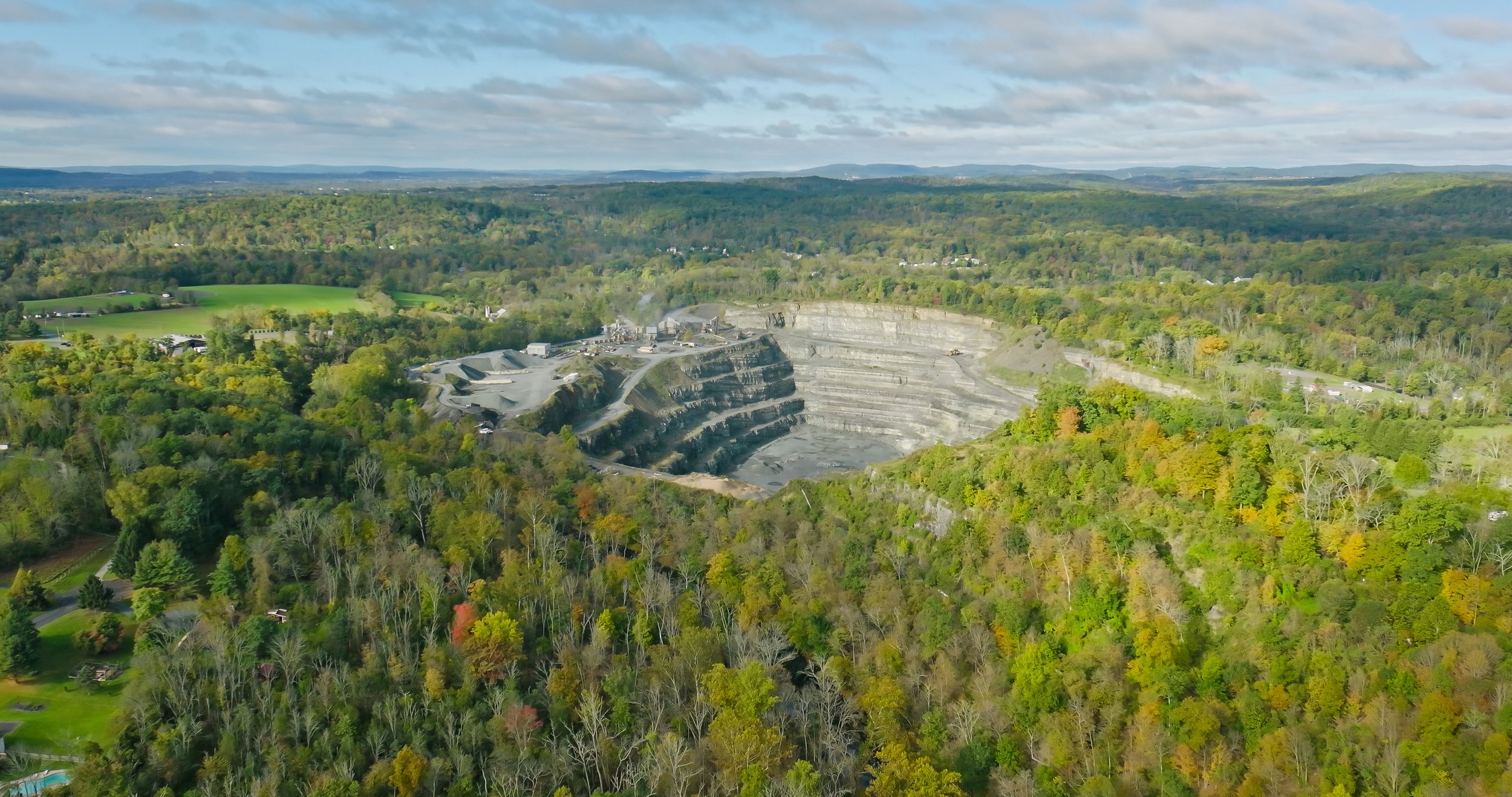 A large open-pit quarry carved into a hillside surrounded by dense green trees and countryside under a partly cloudy sky.