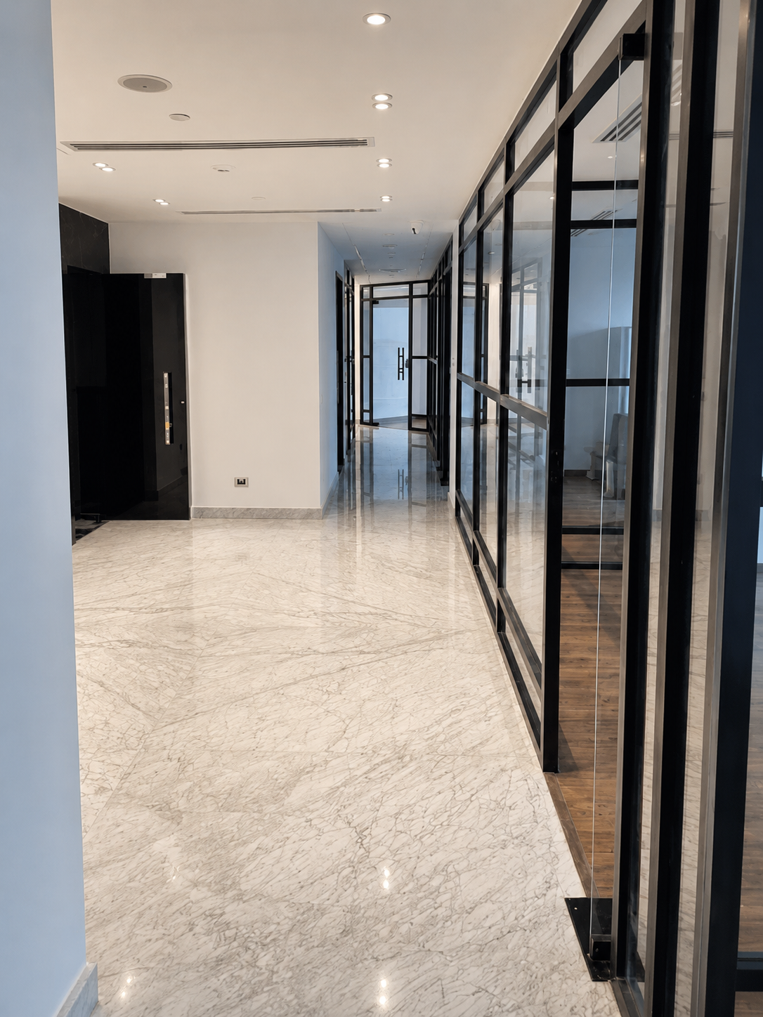Modern hallway with white marble flooring, white ceiling, black-framed glass partitions, and a door at the end.