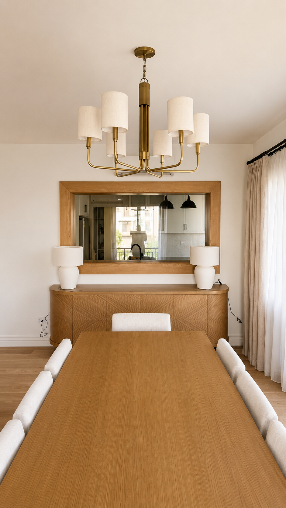 Dining room with a wooden table, white chairs, a large mirror with a wooden frame, two white table lamps, and a gold chandelier with white shades.