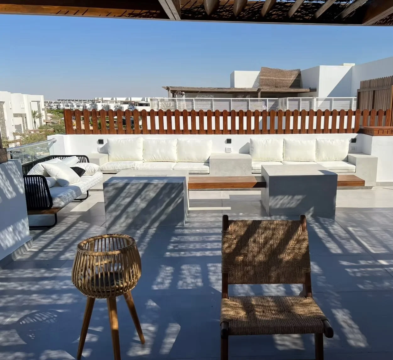 Rooftop patio with white sofa and chairs, wooden fencing, and outdoor furniture, with shadows cast by pergola on sunny day.