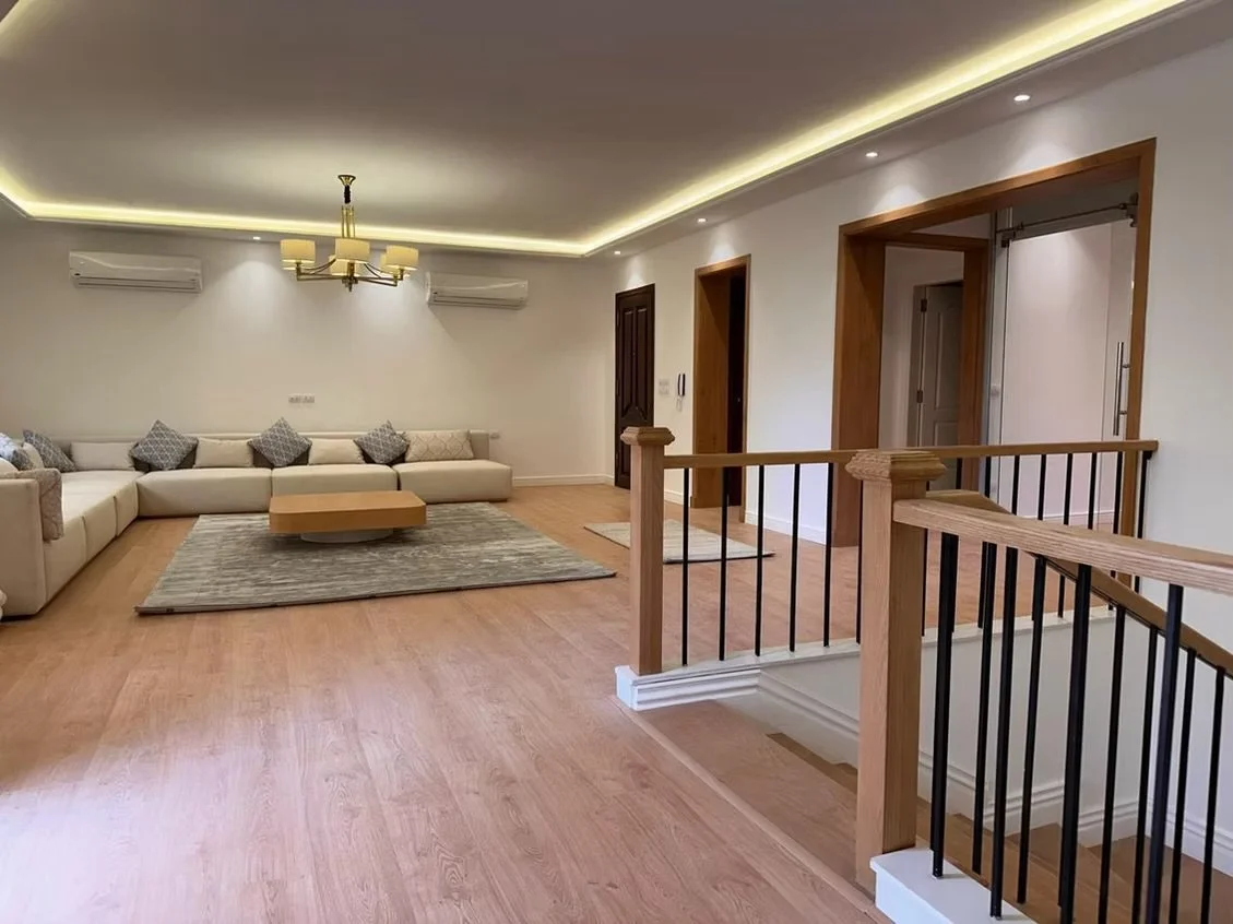 Living room with a large beige sectional sofa, a wooden coffee table, and a gray area rug, with wall-mounted air conditioning units, a chandelier, and recessed lighting.