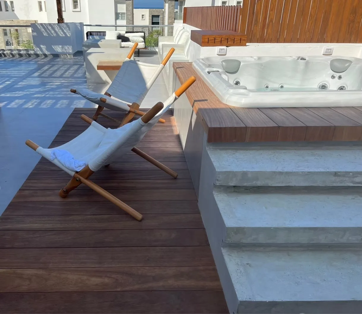 Outdoor rooftop patio with two deck chairs, a hot tub, and wooden flooring with a city view in the background.