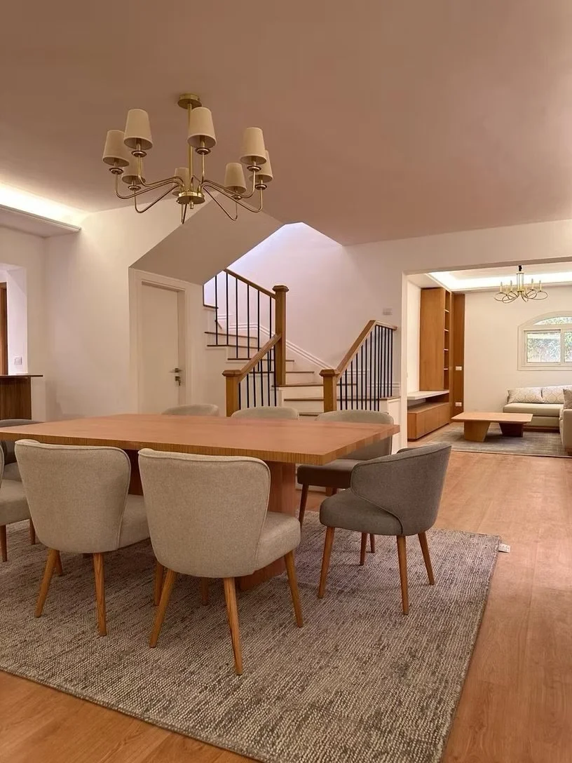 Interior of a dining area with a wooden table and six upholstered chairs on a textured rug, a staircase with wooden and metal railing, and a living room with a TV console, built-in shelves, and a sofa near a window with a chandelier hanging from the ceiling.