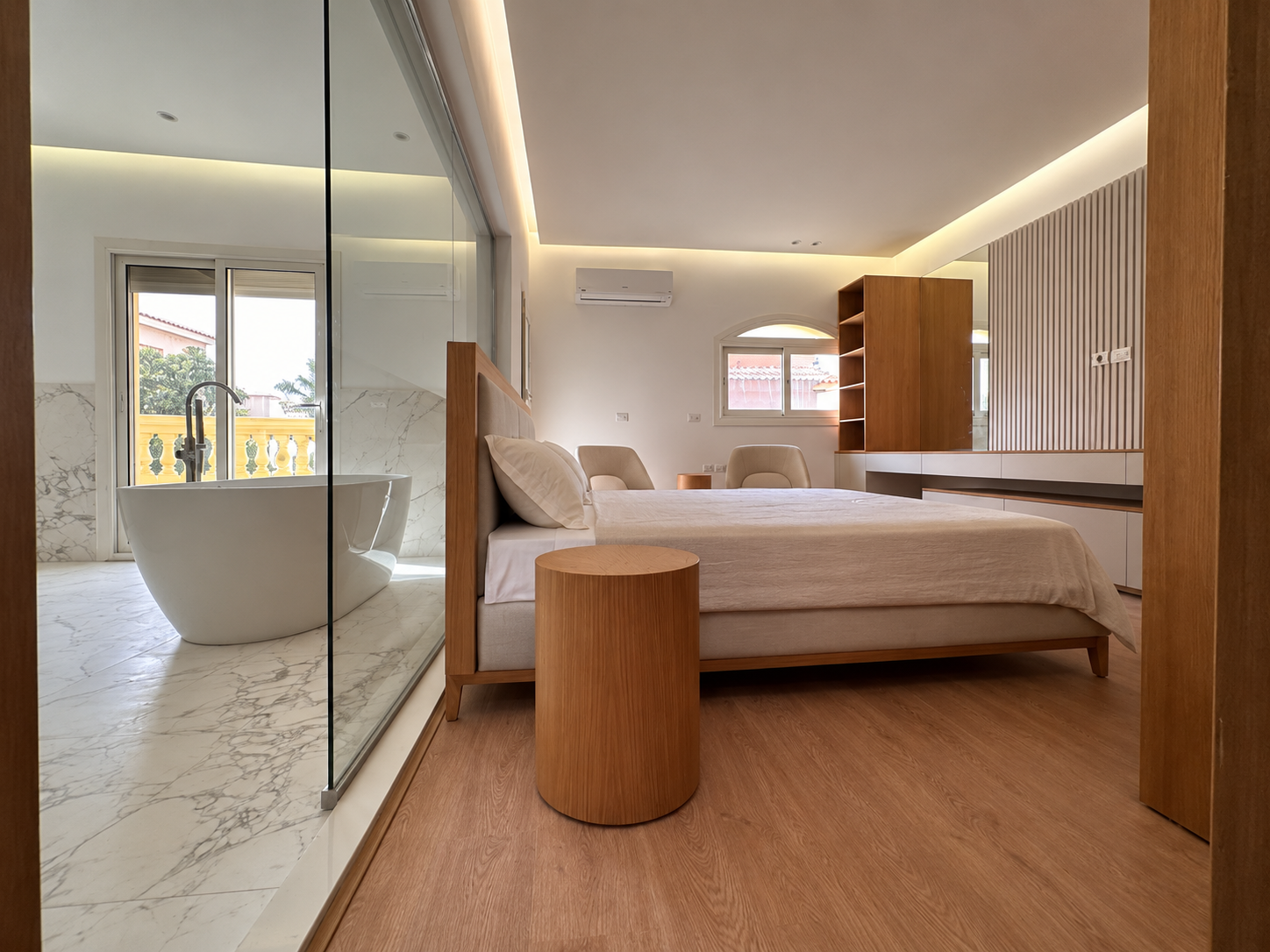 Modern bedroom with a wooden bed frame, white bedding, and a round wooden side table. An interior wall features wooden slats and built-in white drawers. There is a large window and a smaller window, with a balcony outside. Adjacent, a marble bathroom with a freestanding bathtub is visible through a glass partition.
