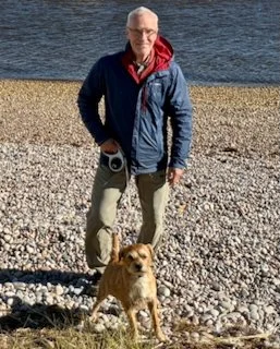 An older man standing on a rocky beach with a small dog at his feet, with water in the background.