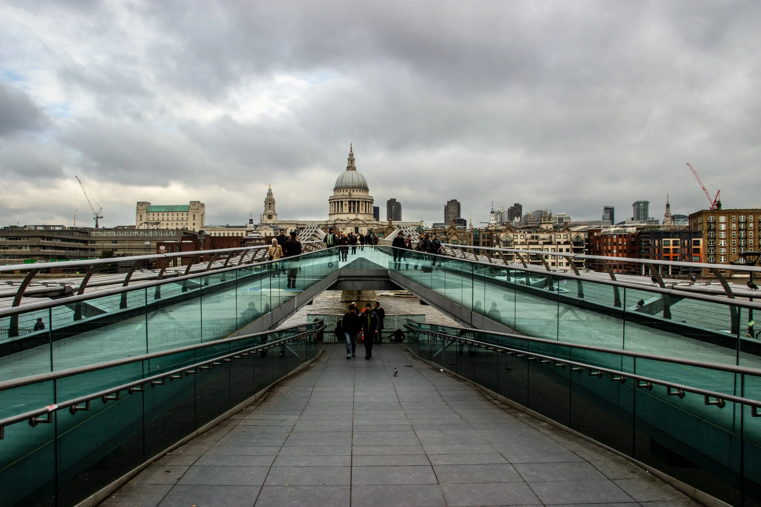 View of St. Paul’s Cathedral in London from Millennium Bridge, a modern pedestrian walkway with glass sides and metal railings, under a cloudy sky with people walking across.