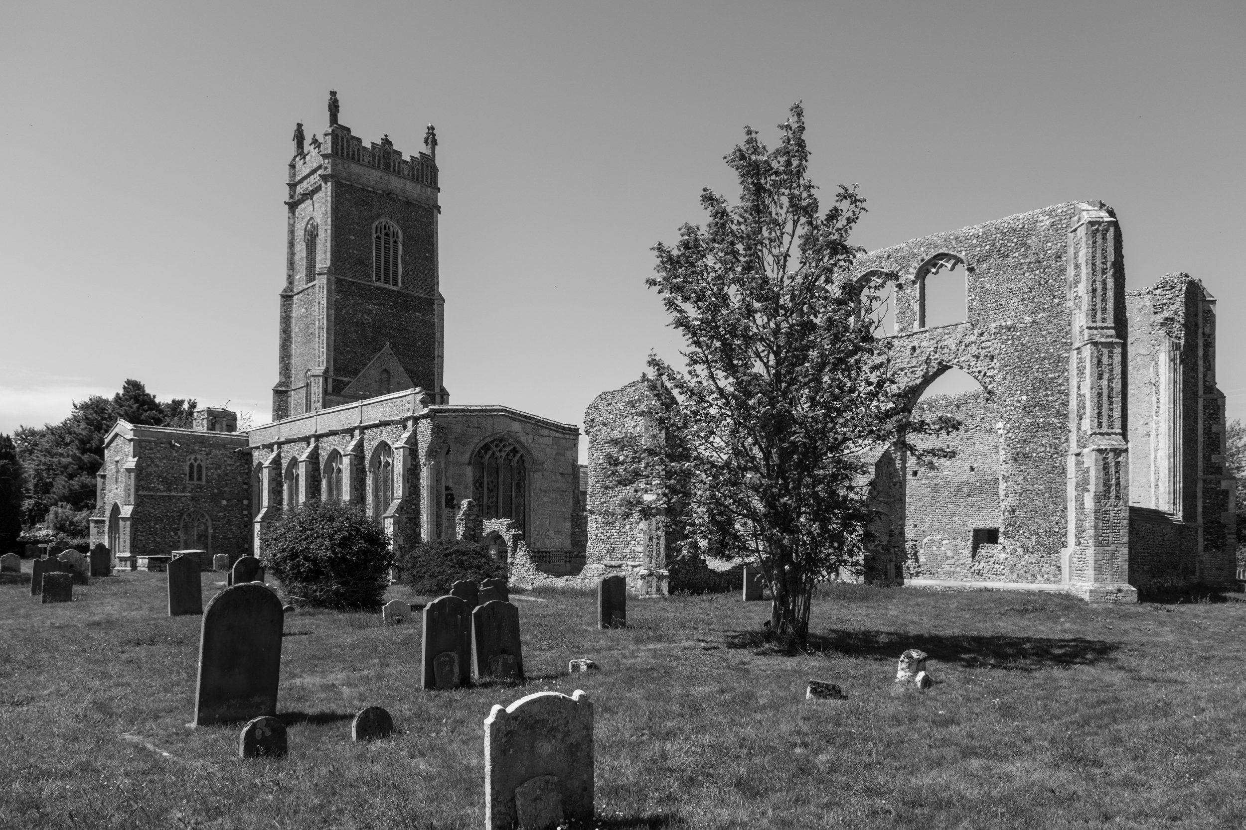 Black and white photograph of an old church with a tall tower, broken walls, and arched windows, surrounded by a graveyard with headstones and a tree in the foreground.