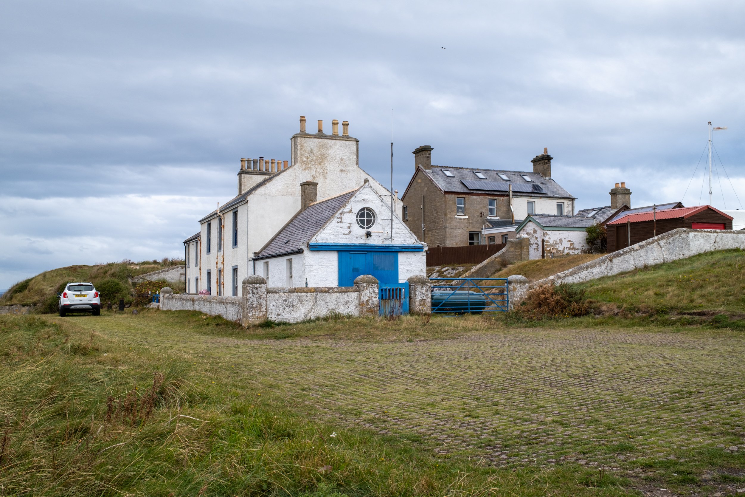 A small white house with a blue door, surrounded by a gray stone fence, with a car parked nearby on a grassy field and other houses in the background under a cloudy sky.