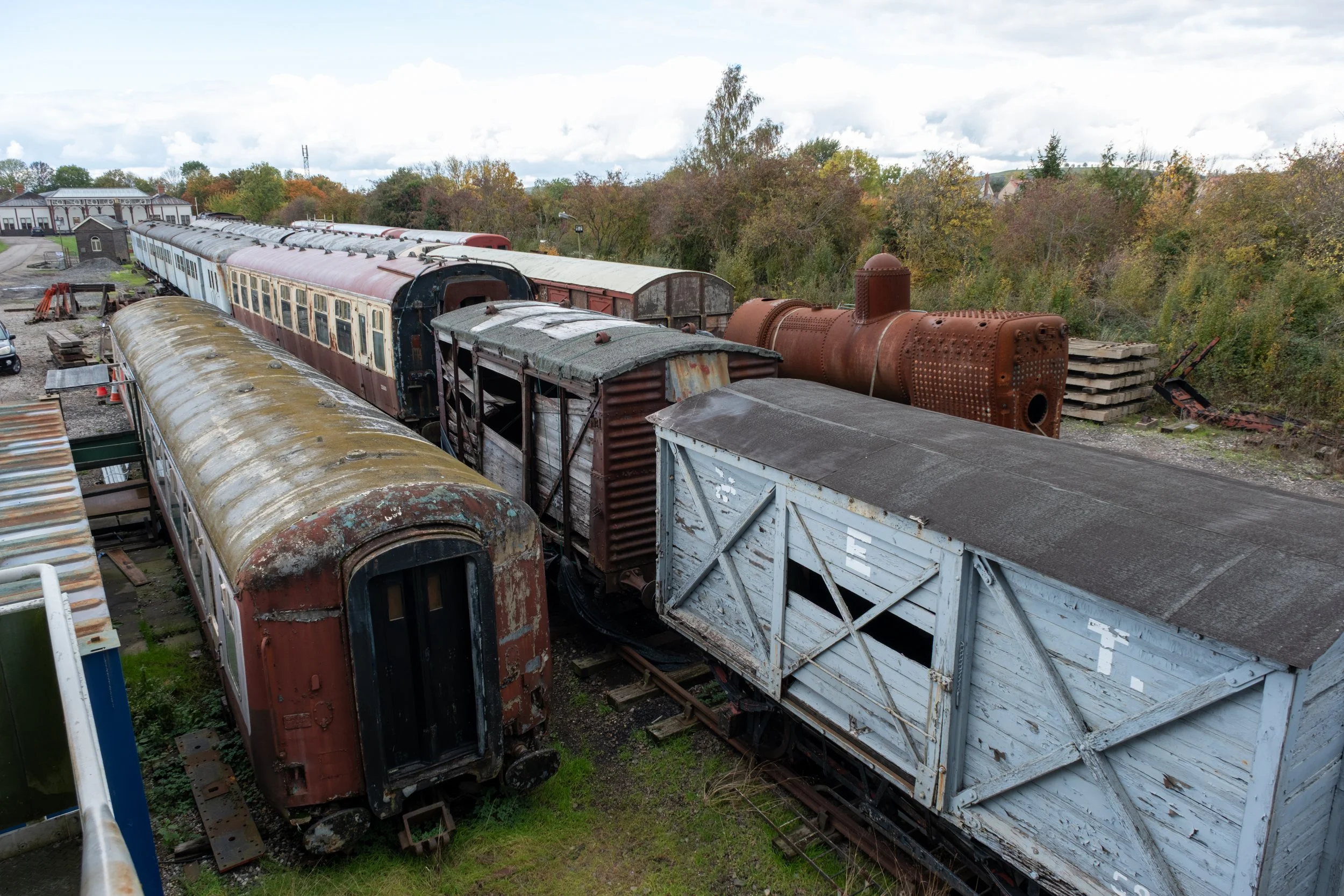 Old, rusty train cars parked on railroad tracks with trees and a building in the background.