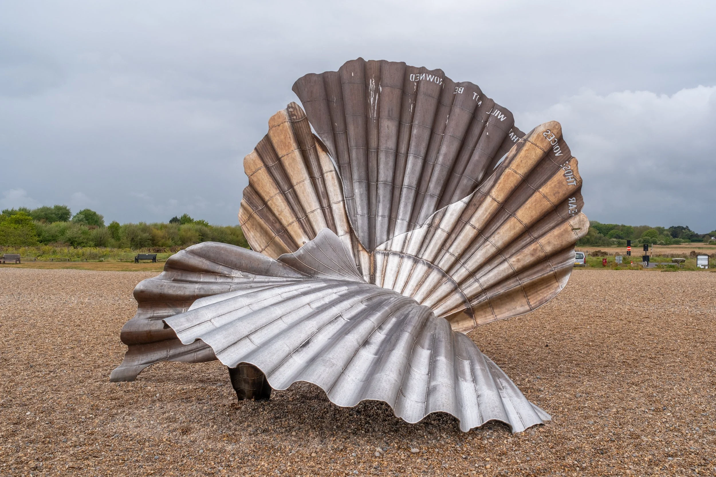 A large metallic sculpture resembling a sea shell outdoors in a park, with a gravel ground, trees in the background, and benches in the distance, under cloudy skies.