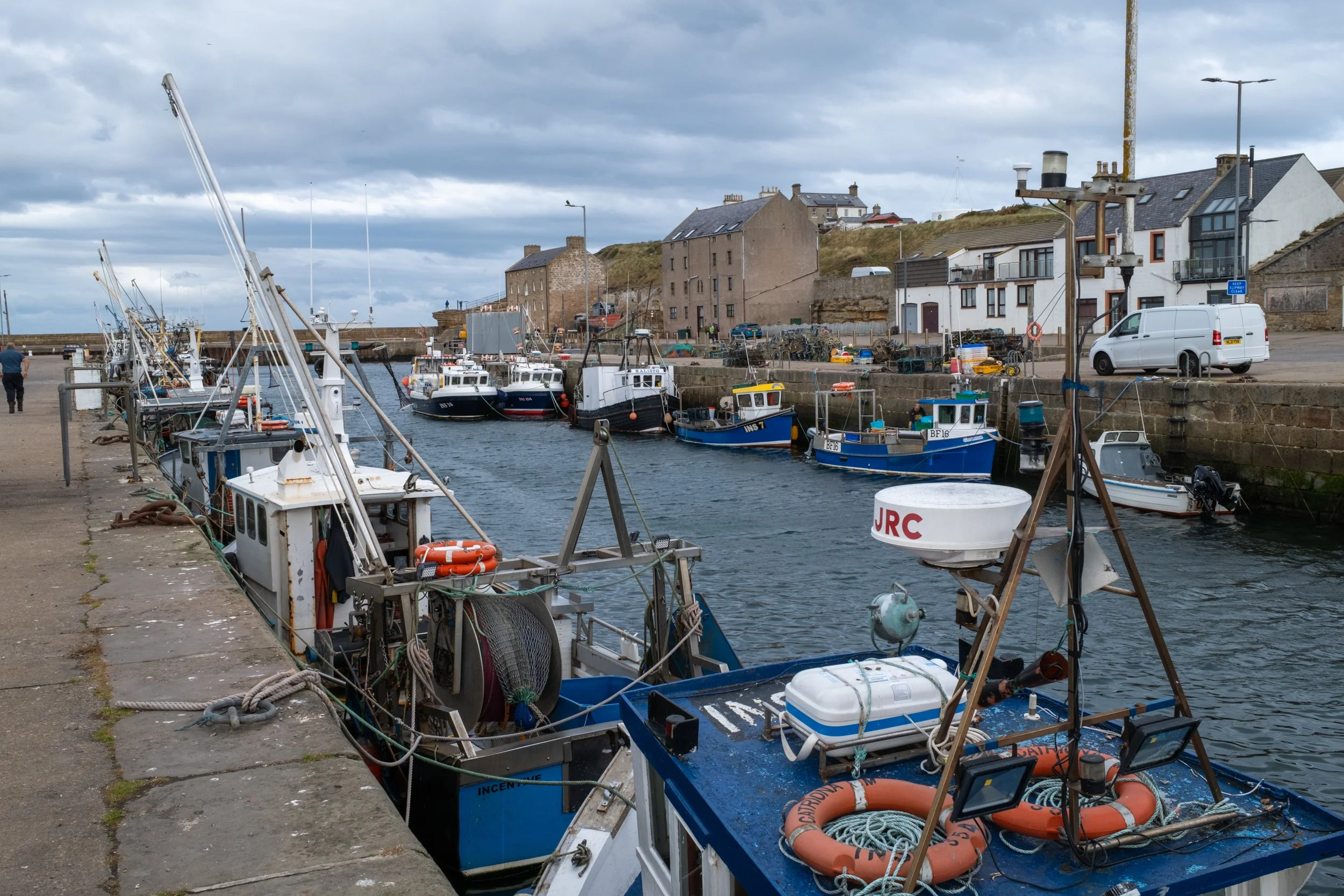 A harbor scene with several boats docked along the pier, buildings on a hillside in the background, and a cloudy sky overhead.