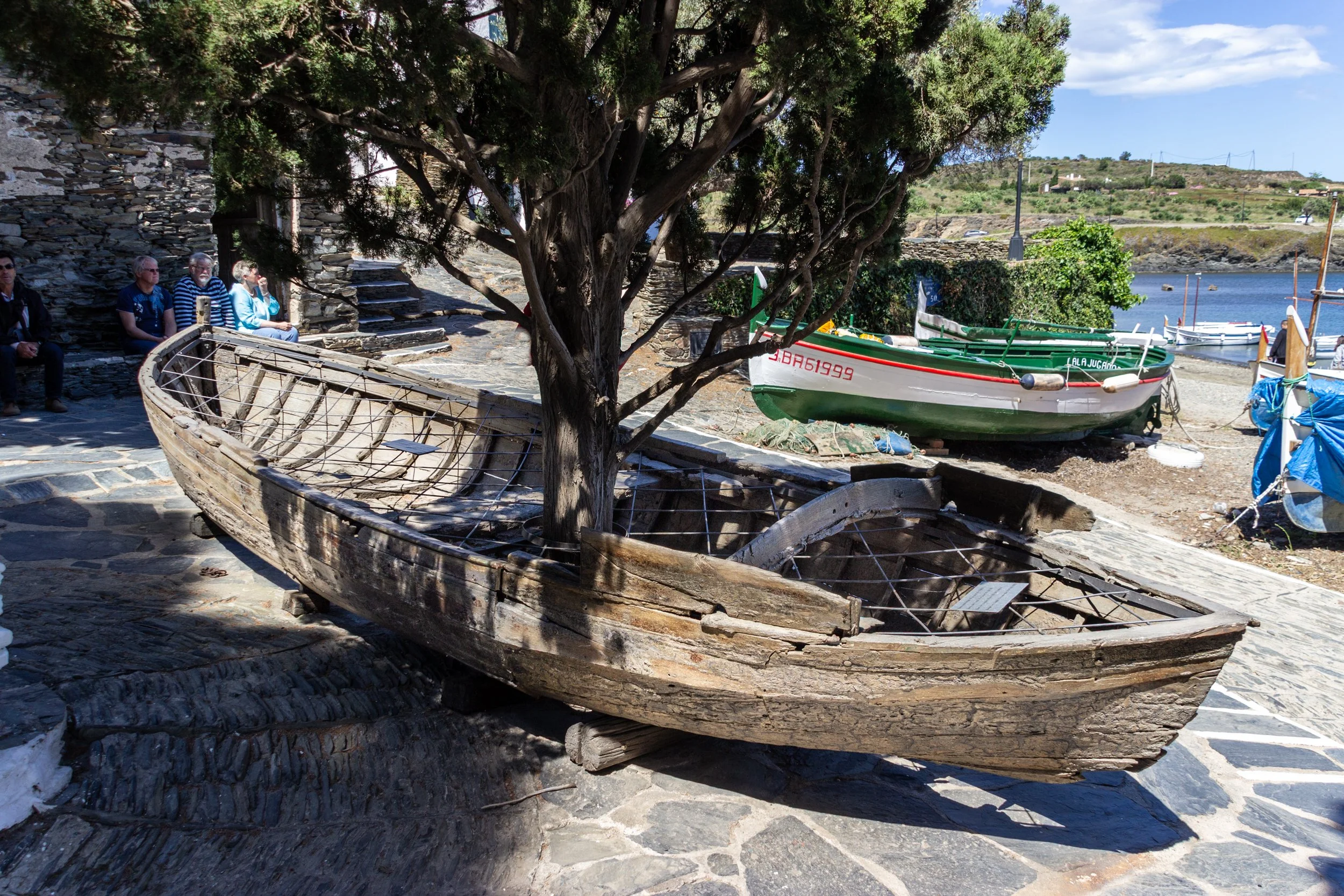 An old wooden boat on land under a large tree, with several people sitting on stone steps nearby. There are other boats visible in the water in the background along a scenic harbor.