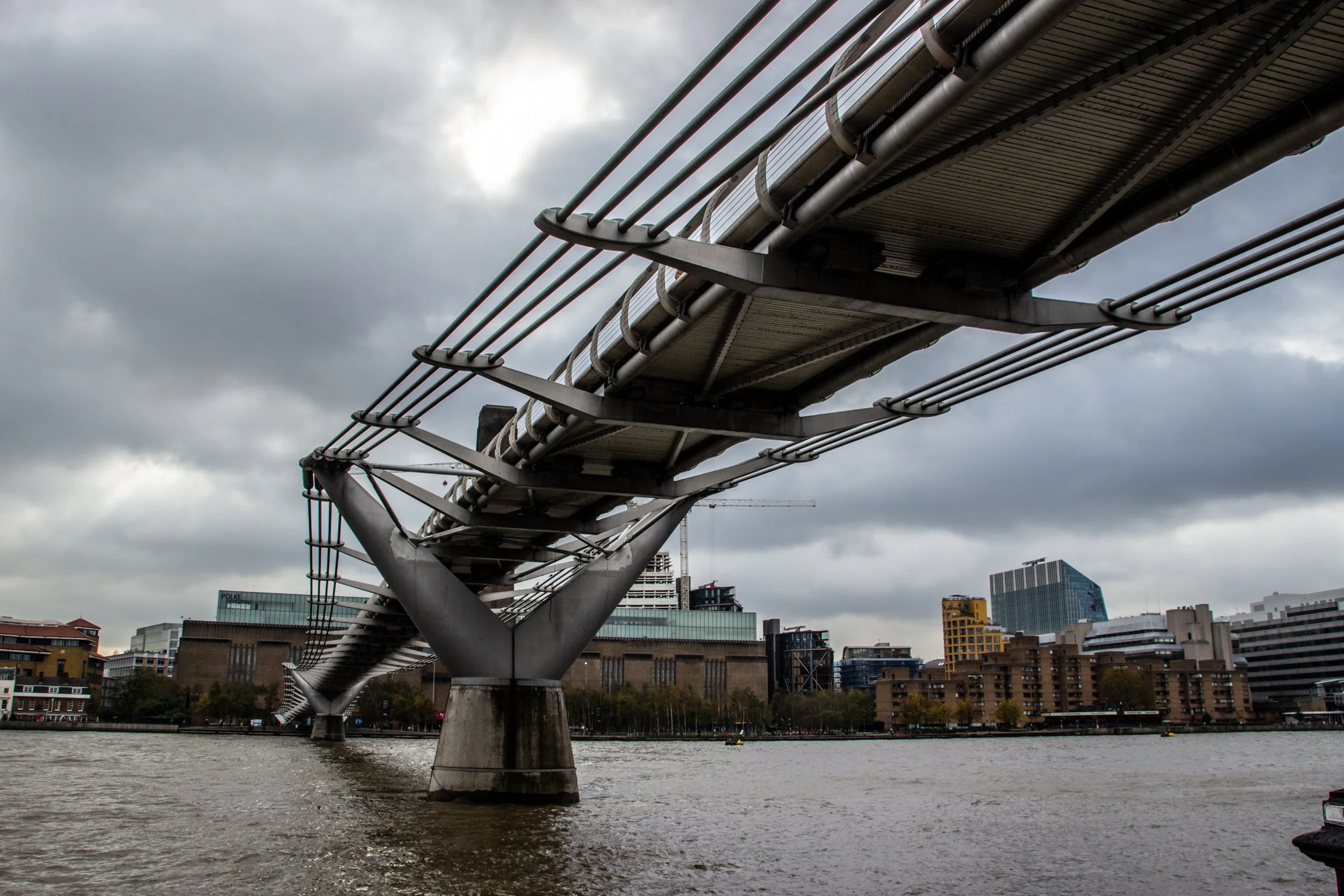 A modern, curved pedestrian bridge over a river in an urban area with tall buildings, under a cloudy sky.