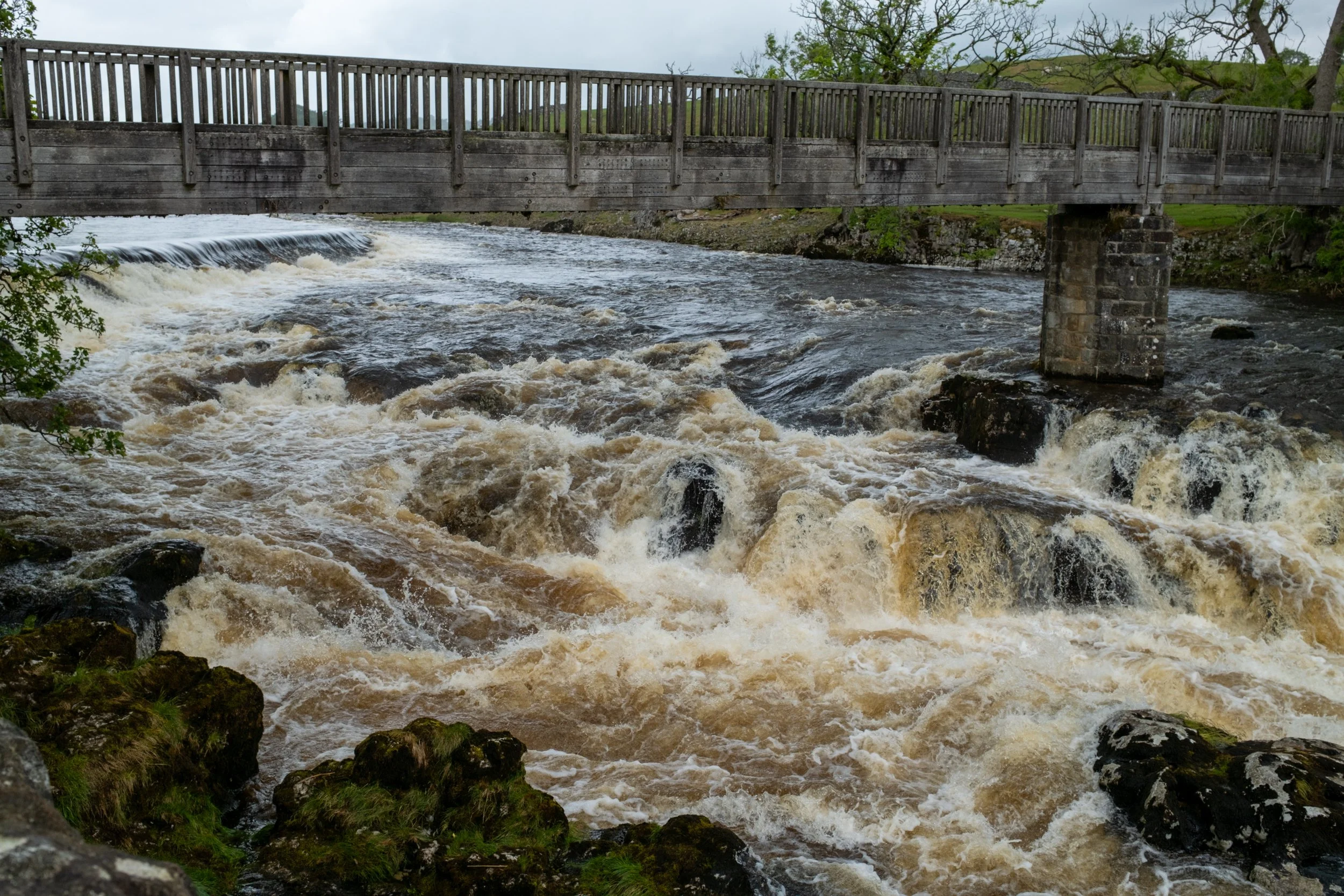 A wooden bridge crossing over a fast-flowing river with whitewater rapids, surrounded by some greenery and trees in the background.