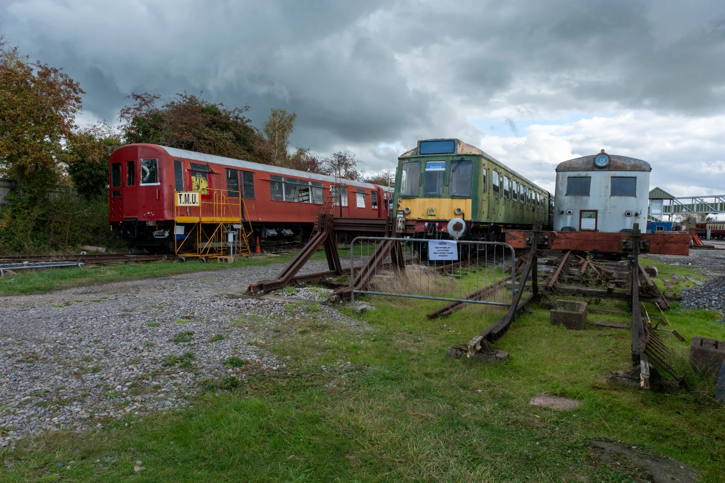 Three old, abandoned trains on a railway track under a cloudy sky. The trains are in a yard with overgrown grass and scattered railway parts.