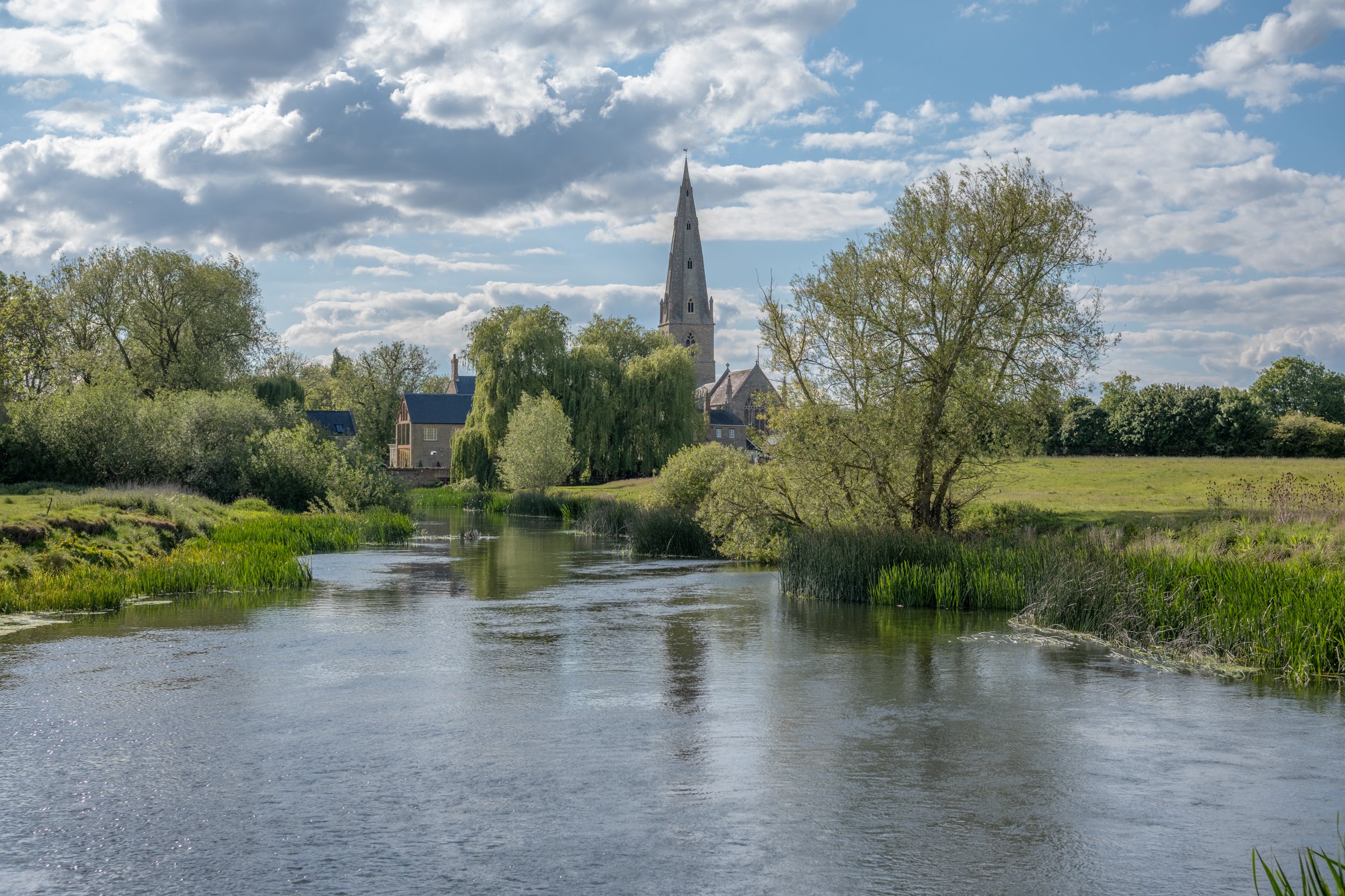 A river flowing through a lush green landscape with trees and houses, and a church with a tall steeple in the background under a partly cloudy sky.