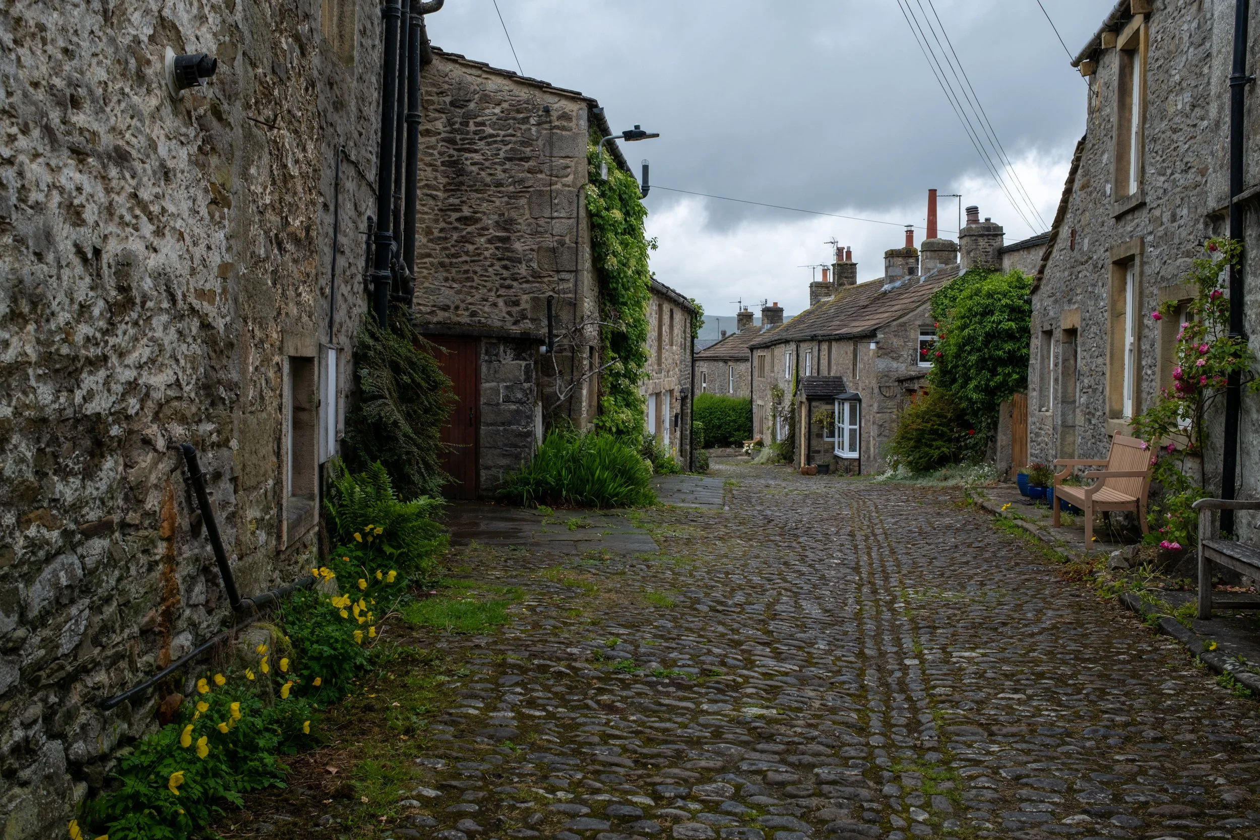 A cobblestone alleyway in a small historic village, with stone houses, greenery, and a bench, under a cloudy sky.