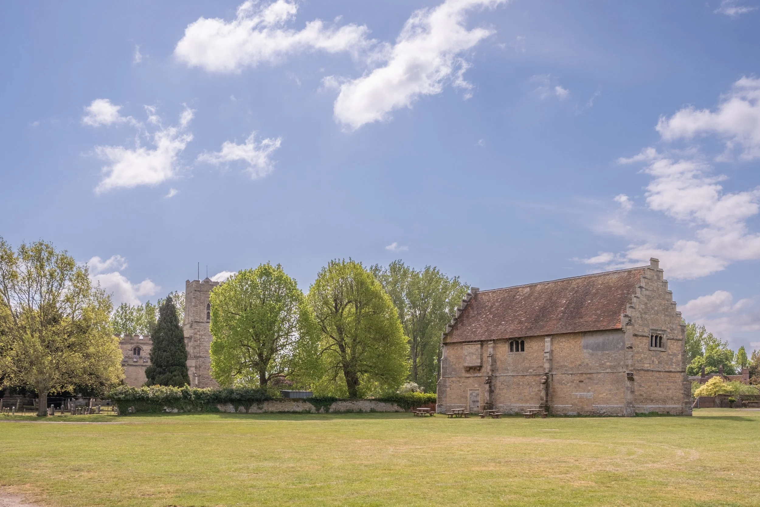 A historic stone church with a steep roof, surrounded by green trees and a grassy field under a partly cloudy sky.