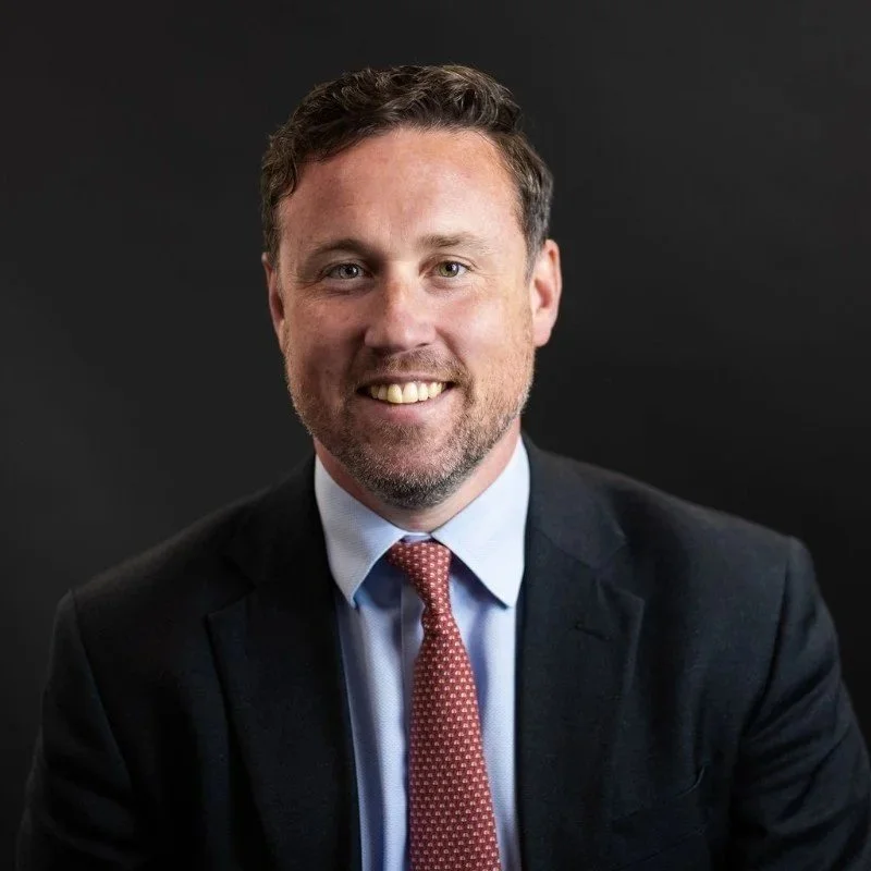 A man with light brown hair, a beard, and a suit with a blue shirt and red polka dot tie, smiling against a dark background.