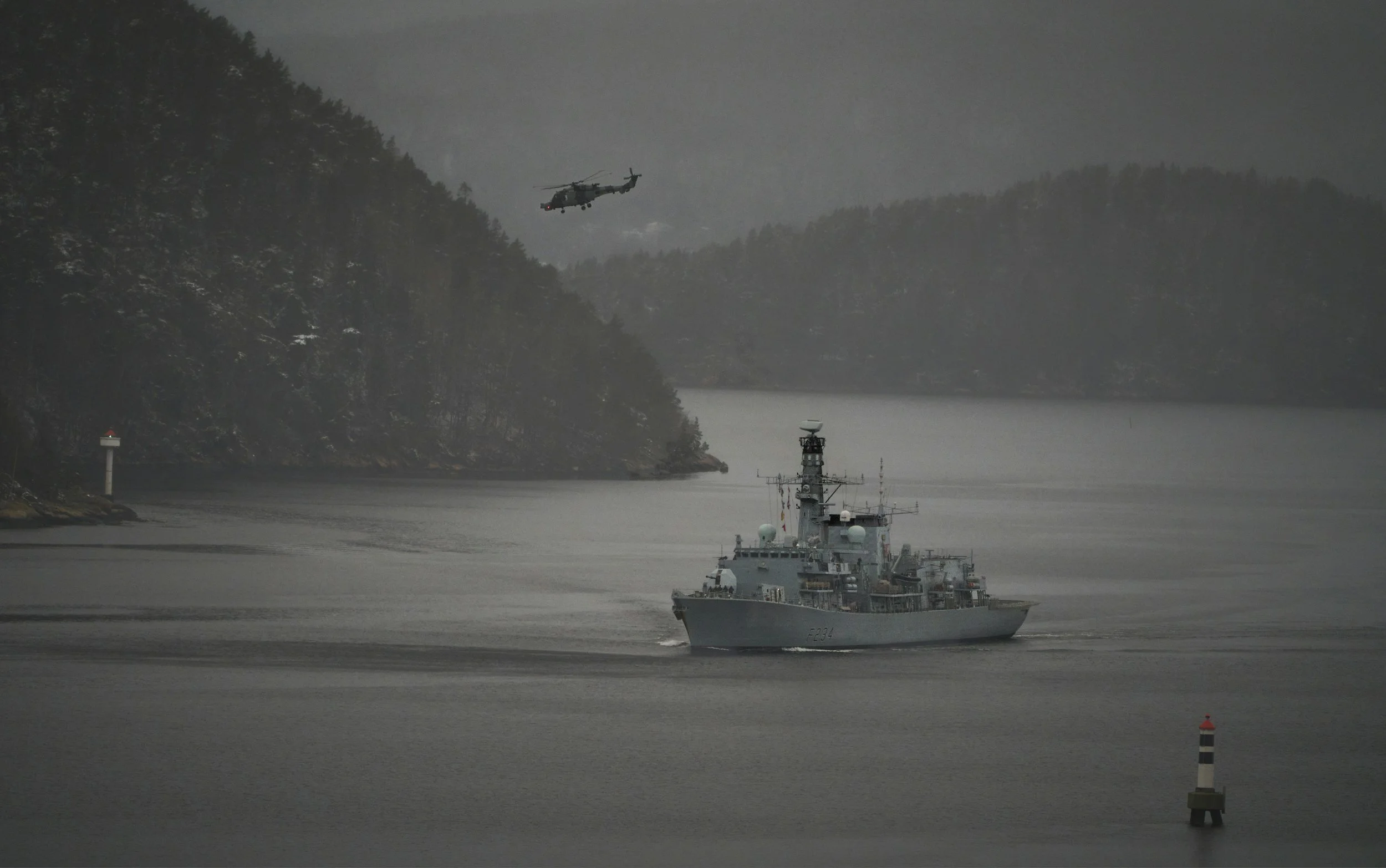 A gray military ship sailing in a river or harbor with a helicopter flying overhead against a backdrop of forested hills and mountains on a cloudy day.