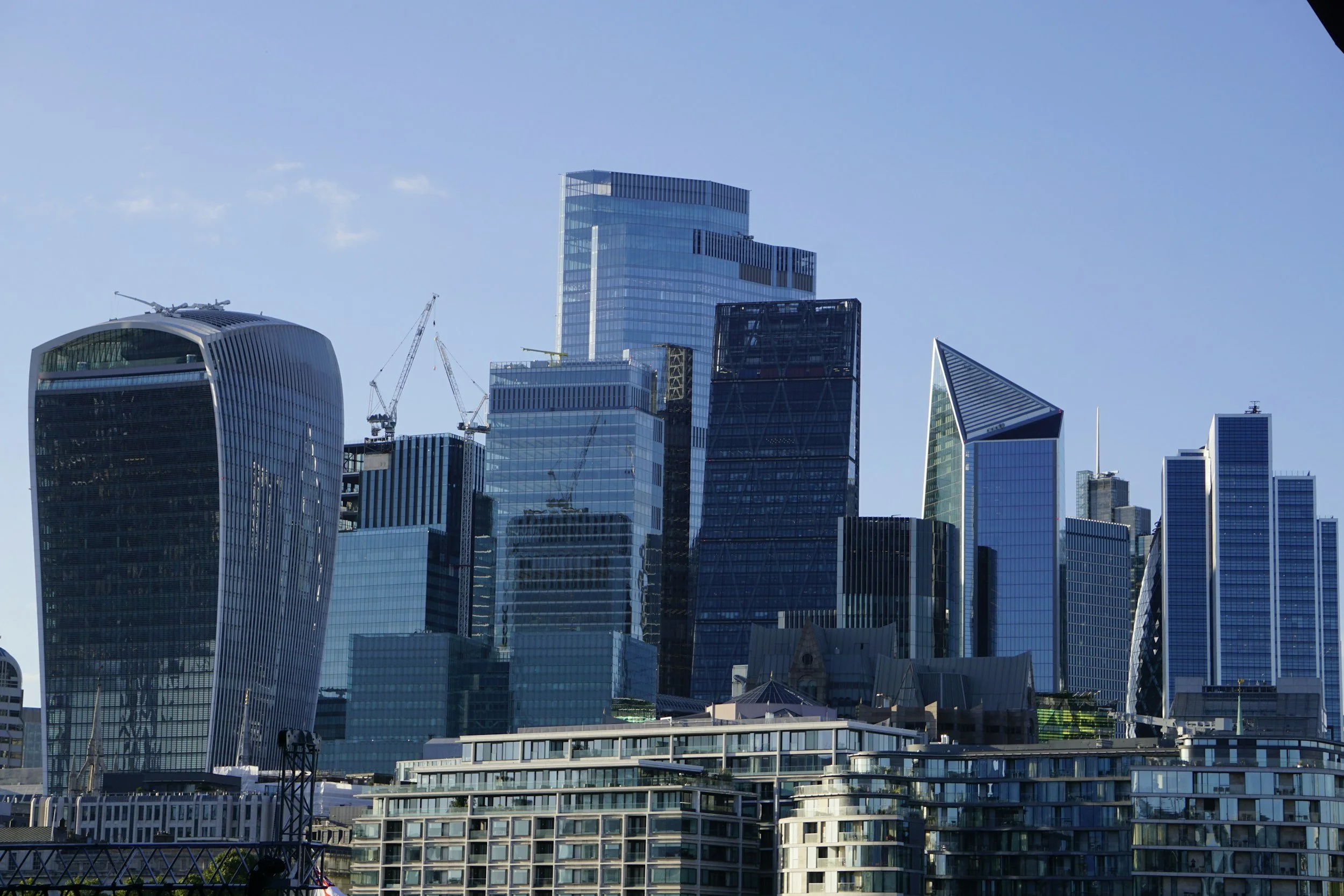 A city skyline with tall modern skyscrapers, some under construction with cranes, against a clear blue sky.