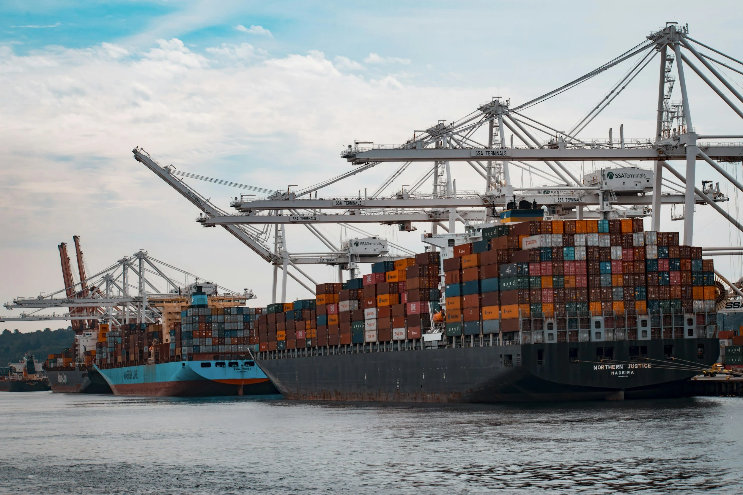 Large cargo ship loaded with stacked shipping containers sailing on a body of water near a port with large white cranes, under a partly cloudy sky.