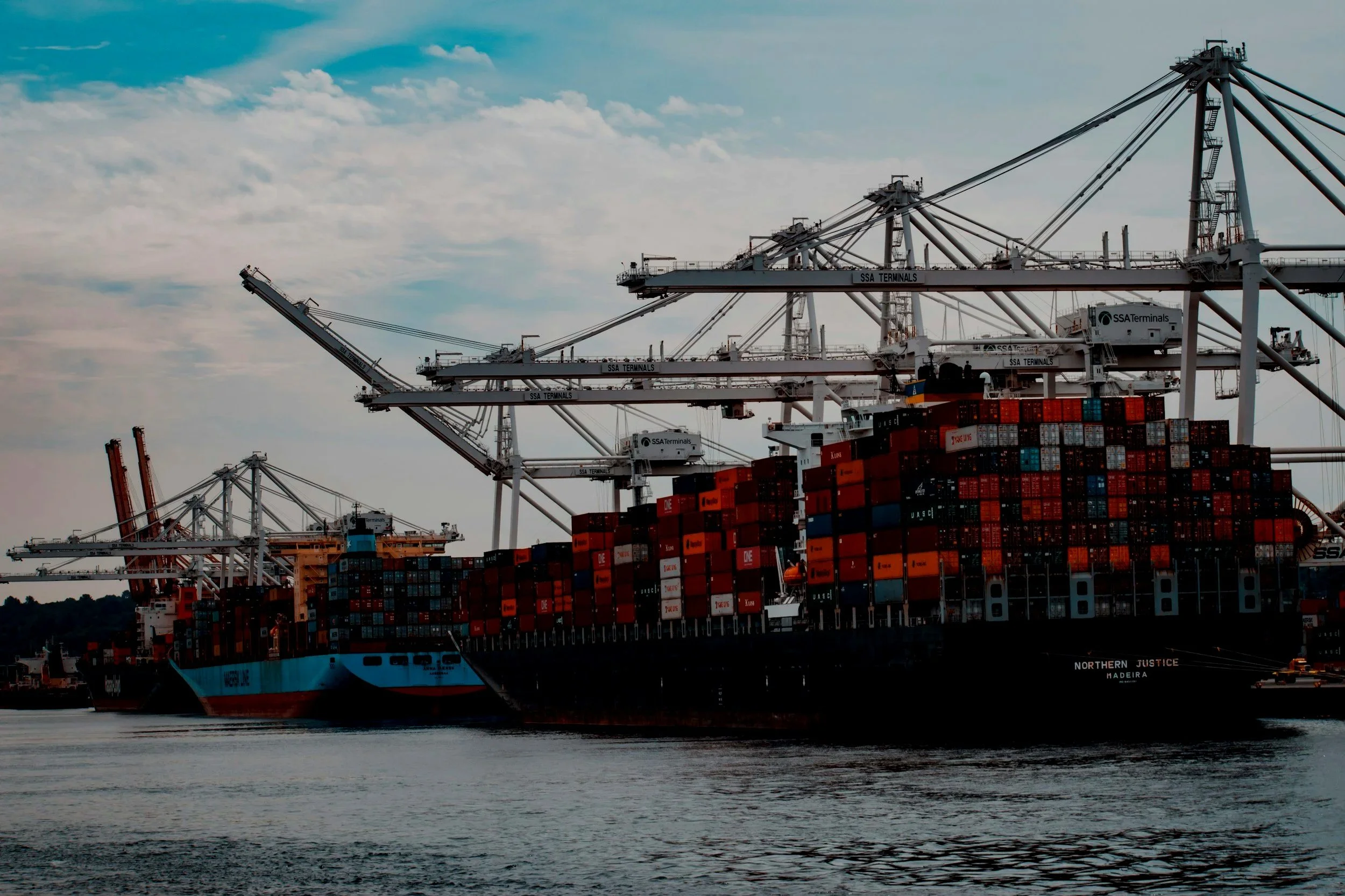 Large cargo ship docked at a port with multiple cranes and stacked shipping containers, under a partly cloudy sky.