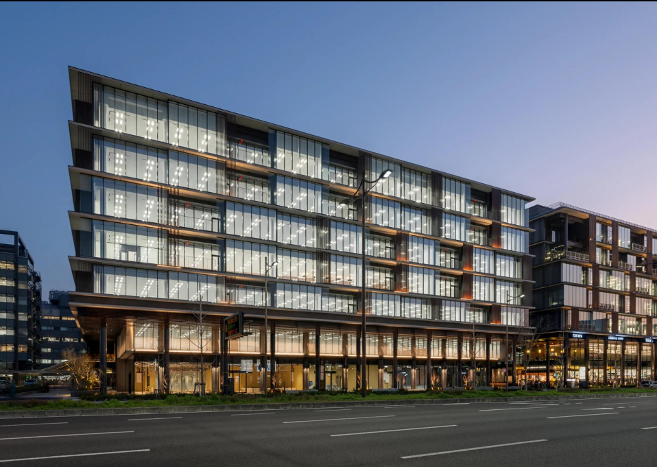 Modern multi-story office building with large glass windows, illuminated interior lights, and an angled facade, located along a city street at dusk.