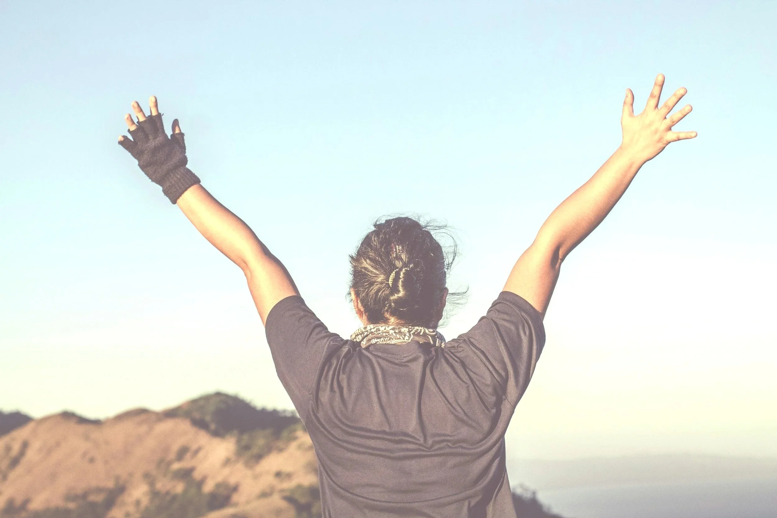 Person from behind raising arms in celebration outdoors with hills and sky in the background.