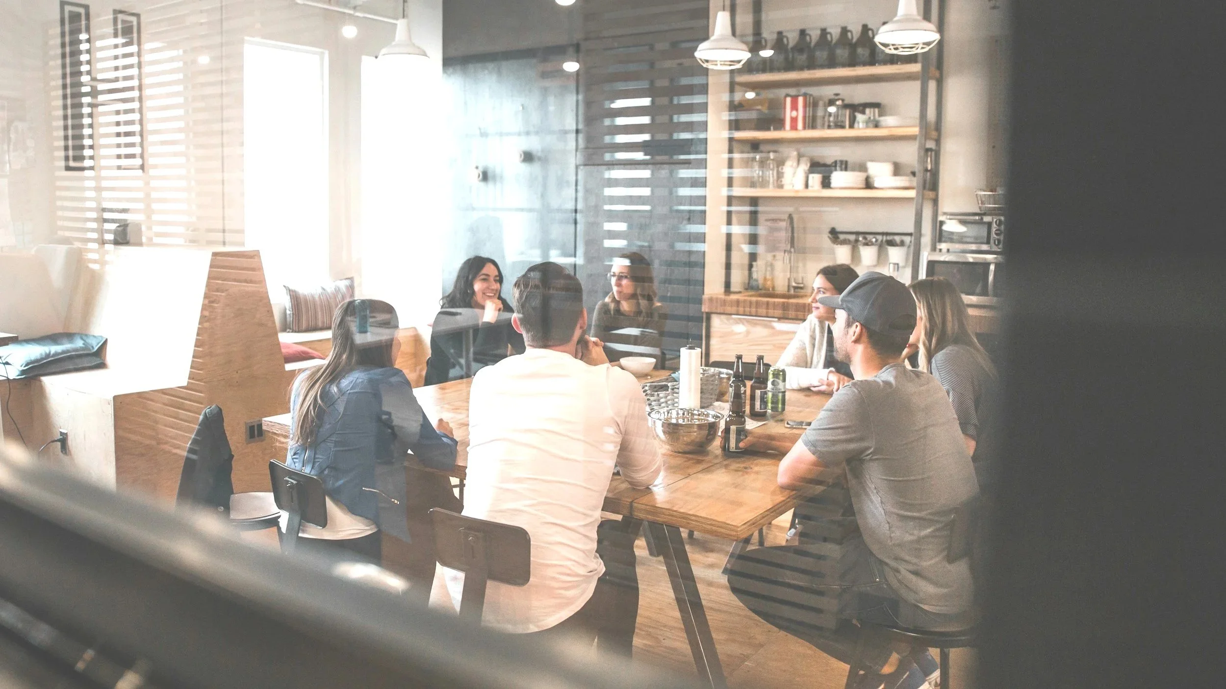 A group of people sitting around a large wooden table in a modern, well-lit cafe or restaurant, engaged in conversation and smiling.