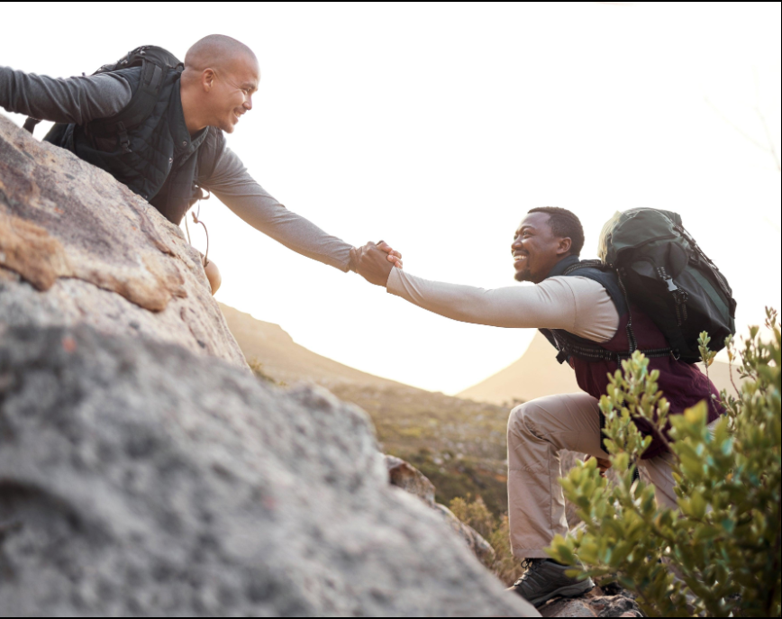 Two men outdoors on a mountain, one reaching down from a higher ledge and holding the other's hand, helping him climb up, both smiling.