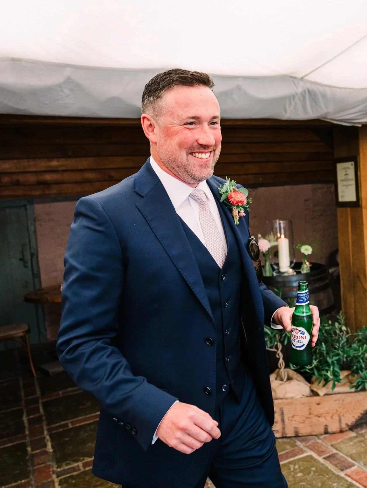 A man in a navy blue suit holding a green Peroni beer bottle, smiling, at a celebration or party indoors with candles and floral decorations in the background.