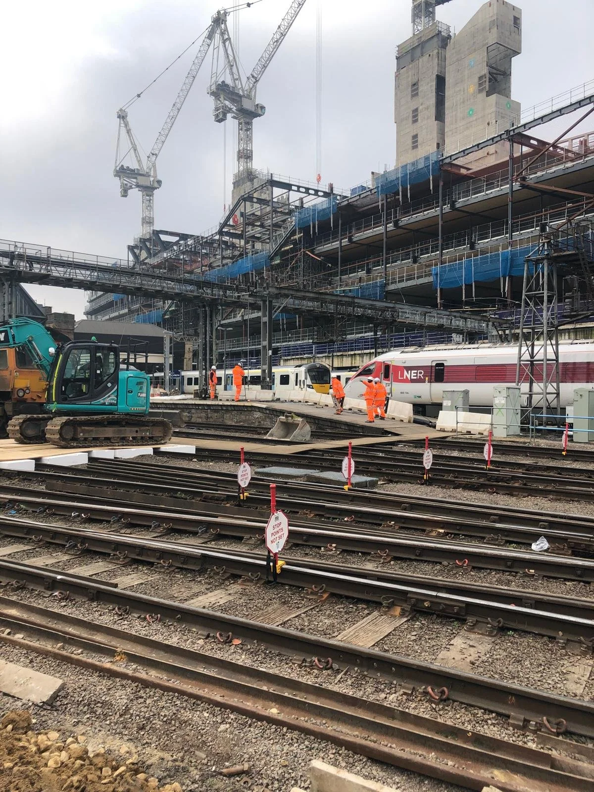 Construction workers in orange safety gear working on train tracks and platform at a railway station under construction. Construction cranes and scaffolding surround the site, with a high-speed train labeled 'LNER' parked nearby. Machinery and safety signs are visible in the area.