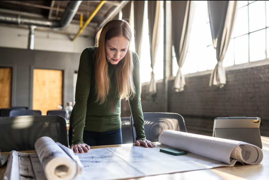 A woman looks over large rolled-out blueprints on a table in a bright industrial-style office.