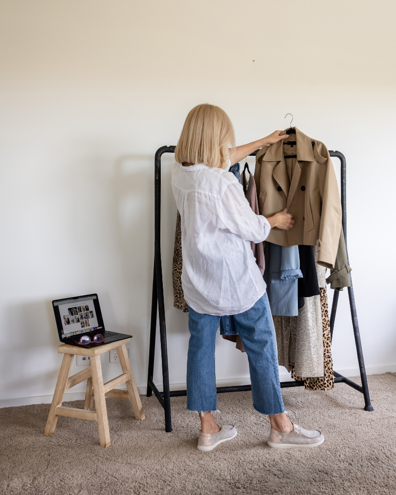 A woman with blonde hair wearing a white shirt and jeans looks at clothing on a black garment rack, with a laptop on a small wooden stool nearby.
