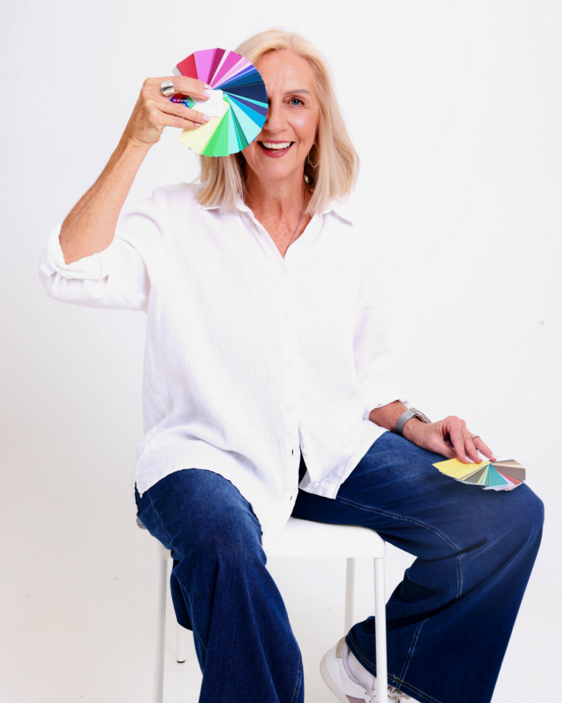 A woman with blonde hair in a white shirt and jeans, smiling and holding a color palette in one hand up to her face, sitting on a white chair against a plain background.