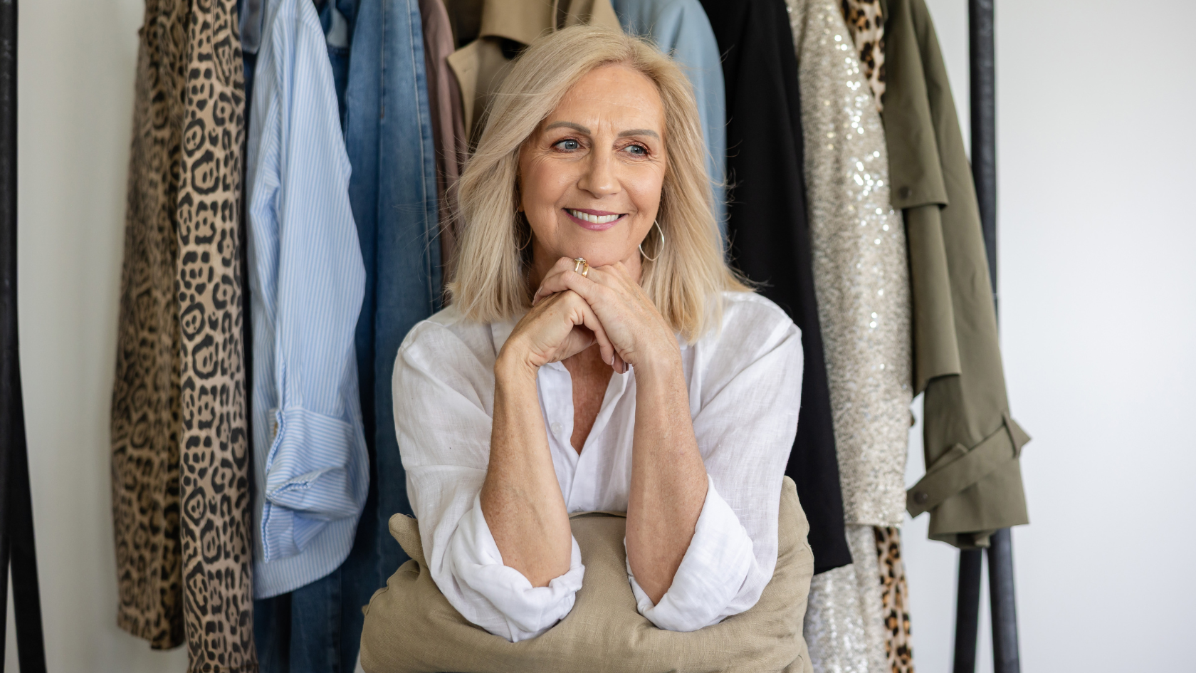 Smiling woman with blonde hair sitting in front of a clothing rack with various jackets and shirts.