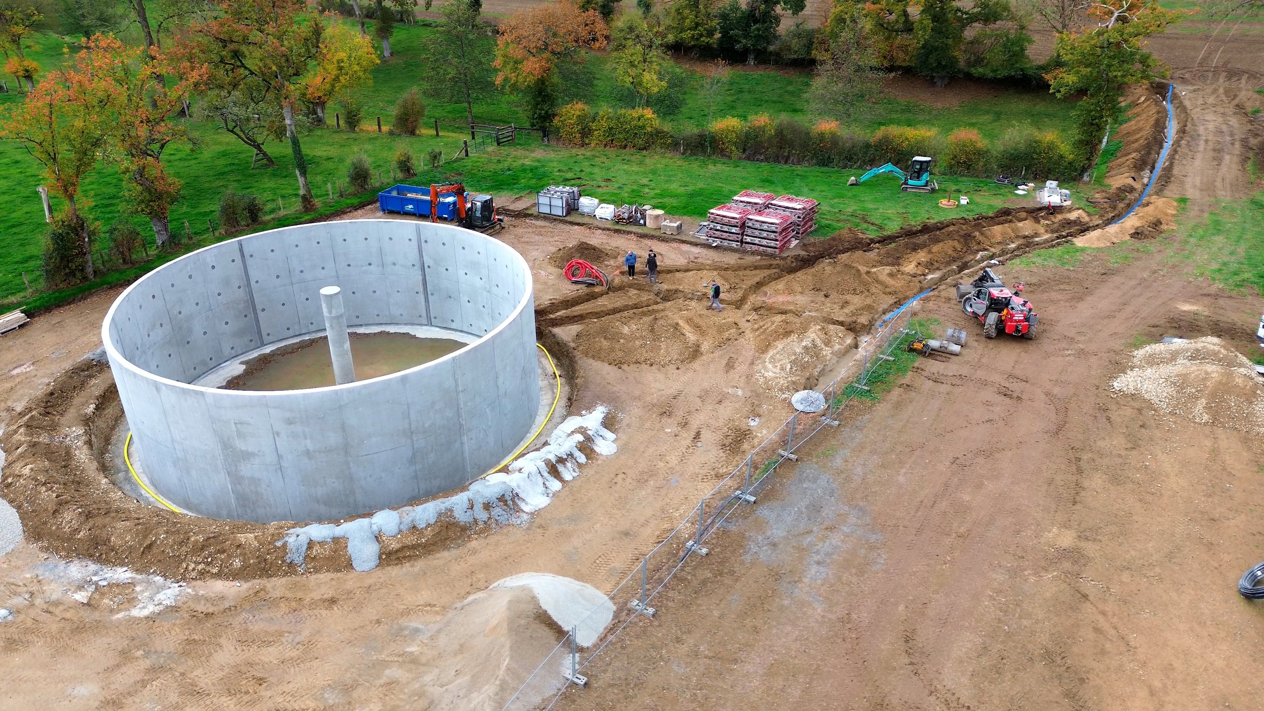 Construction en cours avec un grand réservoir cylindrique en béton, un engin de chantier, et plusieurs ouvriers dans un espace en terre, entouré d'arbres aux feuilles colorées.