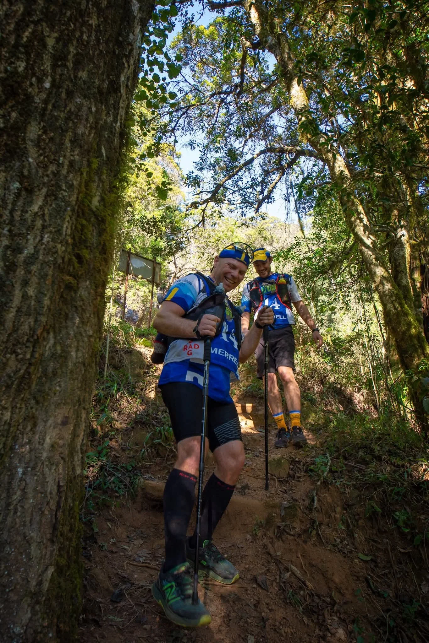 Two smiling men in athletic gear hiking on a forest trail.