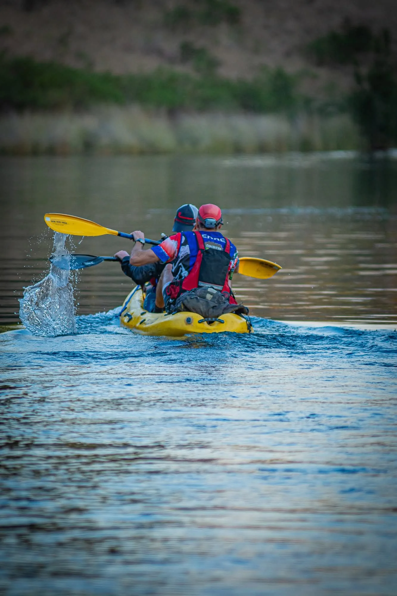 Two people kayaking on a calm river or lake, viewed from behind, with wooded shoreline in the background.