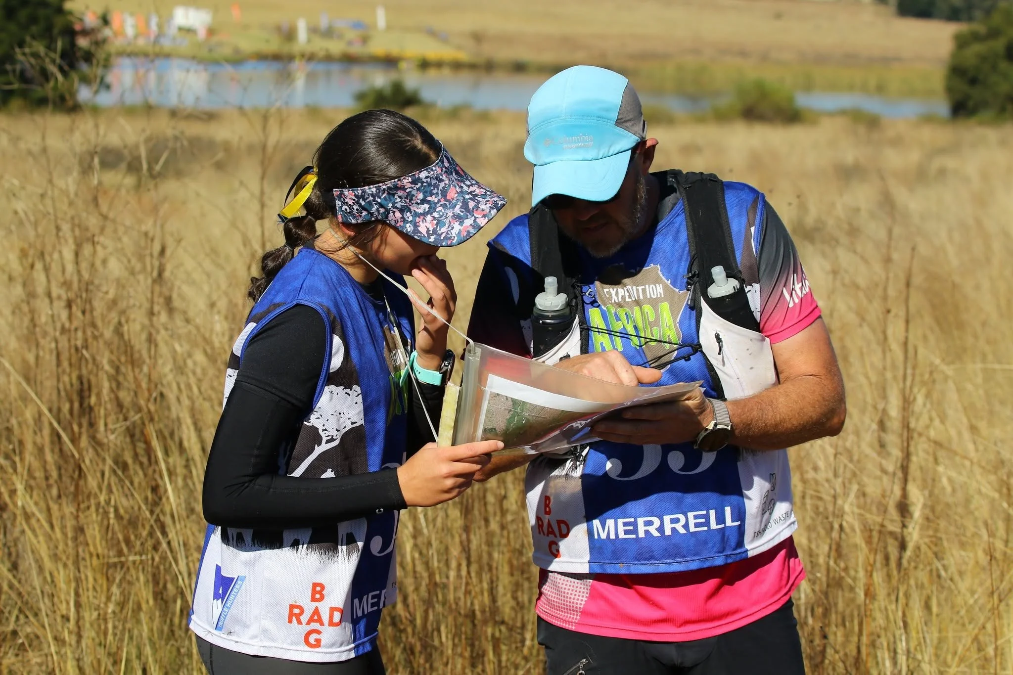Two people in athletic gear look at a map outdoors near tall grass, with a body of water and hills in the background.